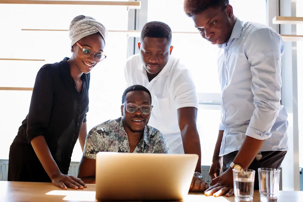 Four colleagues gathered around a laptop, talking and smiling.
